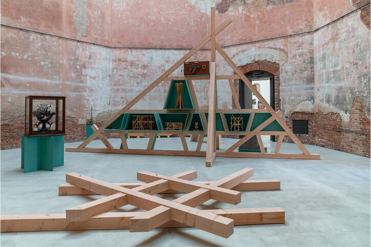 Ein Kunstwerk aus Holzbalken liegt in einem großen Raum mit Ziegelmauerwerk
